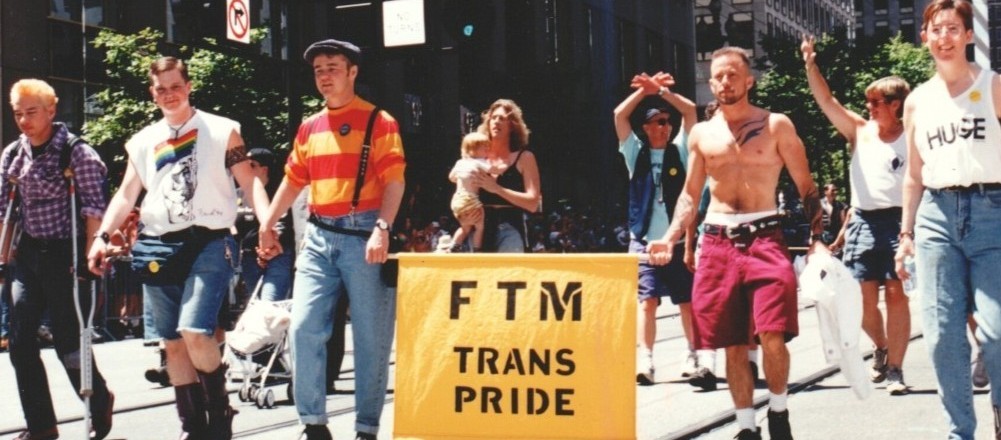 People walking in a parade, five men in front, three people behind them. Two men are carrying a banner that says 'FTM Trans Pride'. Two of the men are holding hands.